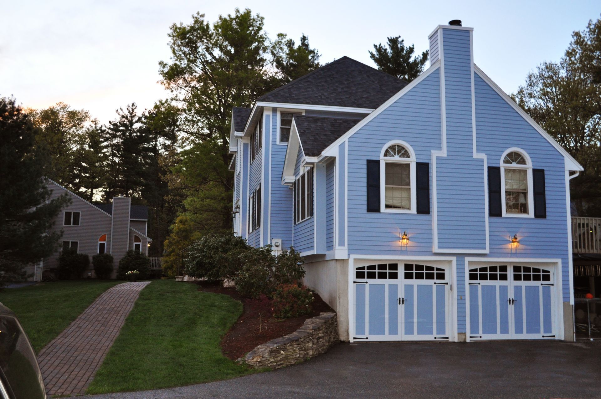 A light blue two-story house with a two-car garage, black shutters, and an arched window, surrounded by trees at dusk.