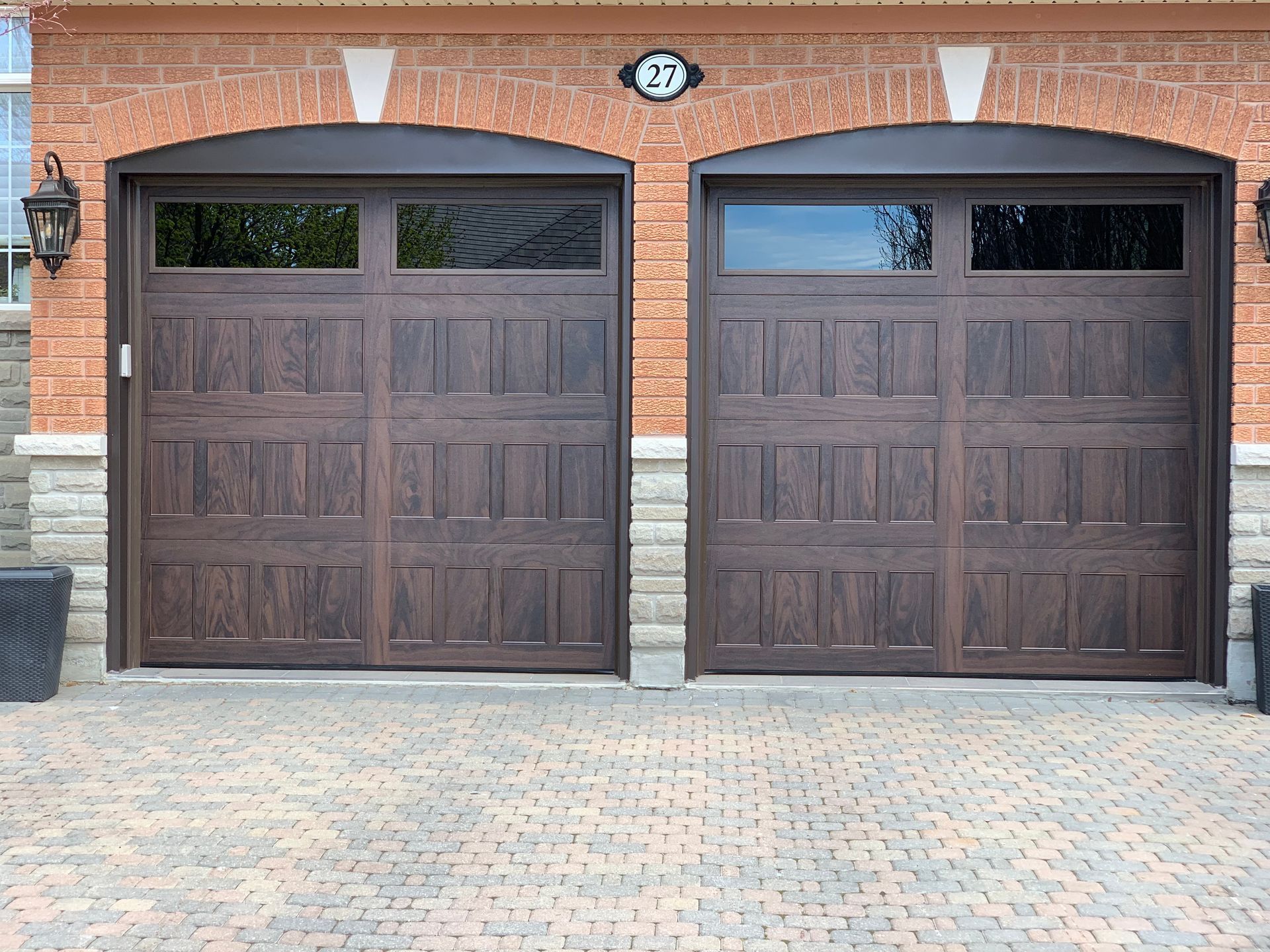 Two dark-wood, multi-paneled arched garage doors with top windows, set in a brick exterior with a stone base.