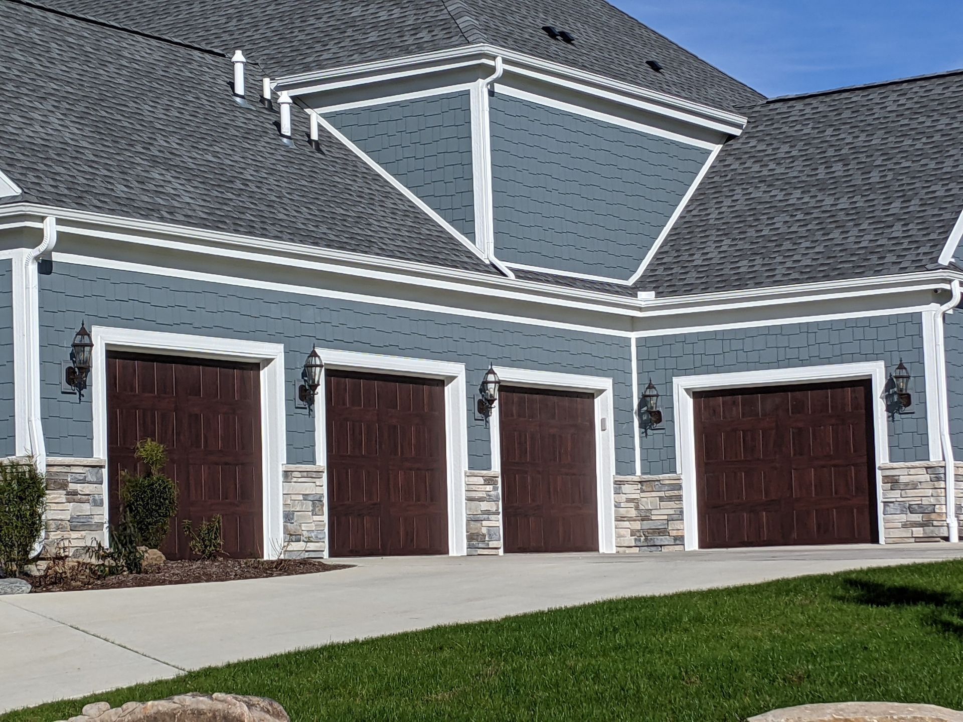 A residential three-car garage with dark brown doors, blue siding, stone base, and gray shingled roof on a sunny day.