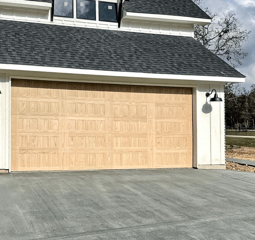 A beige wooden garage door on a white house with a gray shingled roof and a black outdoor light fixture.