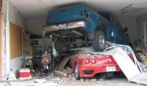 A blue truck crashed on top of a red sports car inside a damaged garage.