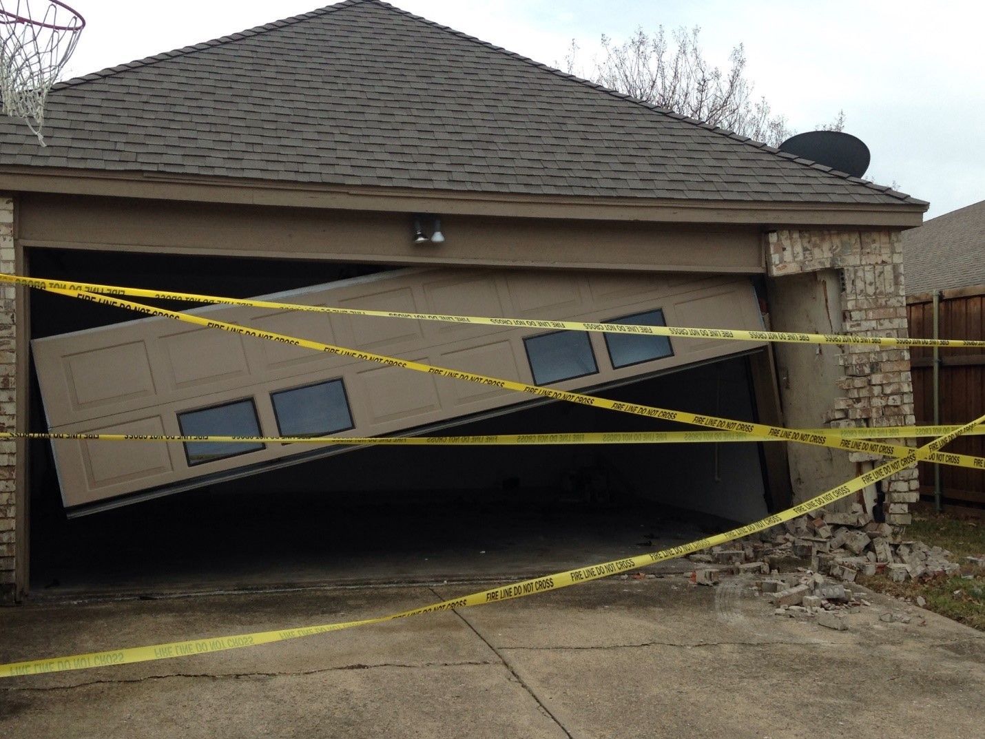Damaged garage with tilted door; caution tape across opening; brickwork damage.
