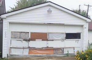 A dilapidated white garage with boarded-up windows and missing panels, in front of a house.
