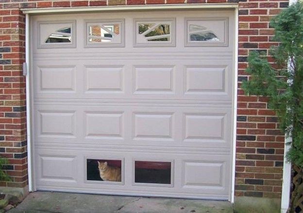 Beige garage door with window panels, cat peering out of lower panel opening. Brick wall.