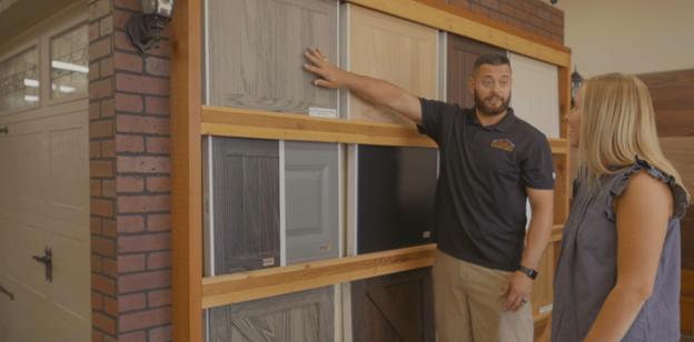 Man pointing at door samples; woman listens. Brick wall & garage visible.