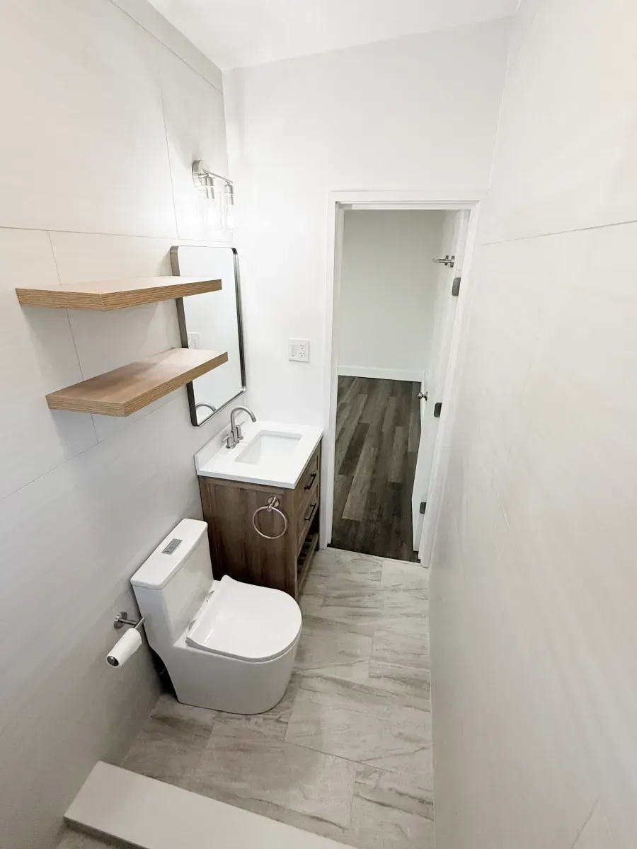 A small modern bathroom with a white toilet, wooden vanity with a white sink, two wooden shelves, and light-colored floors.