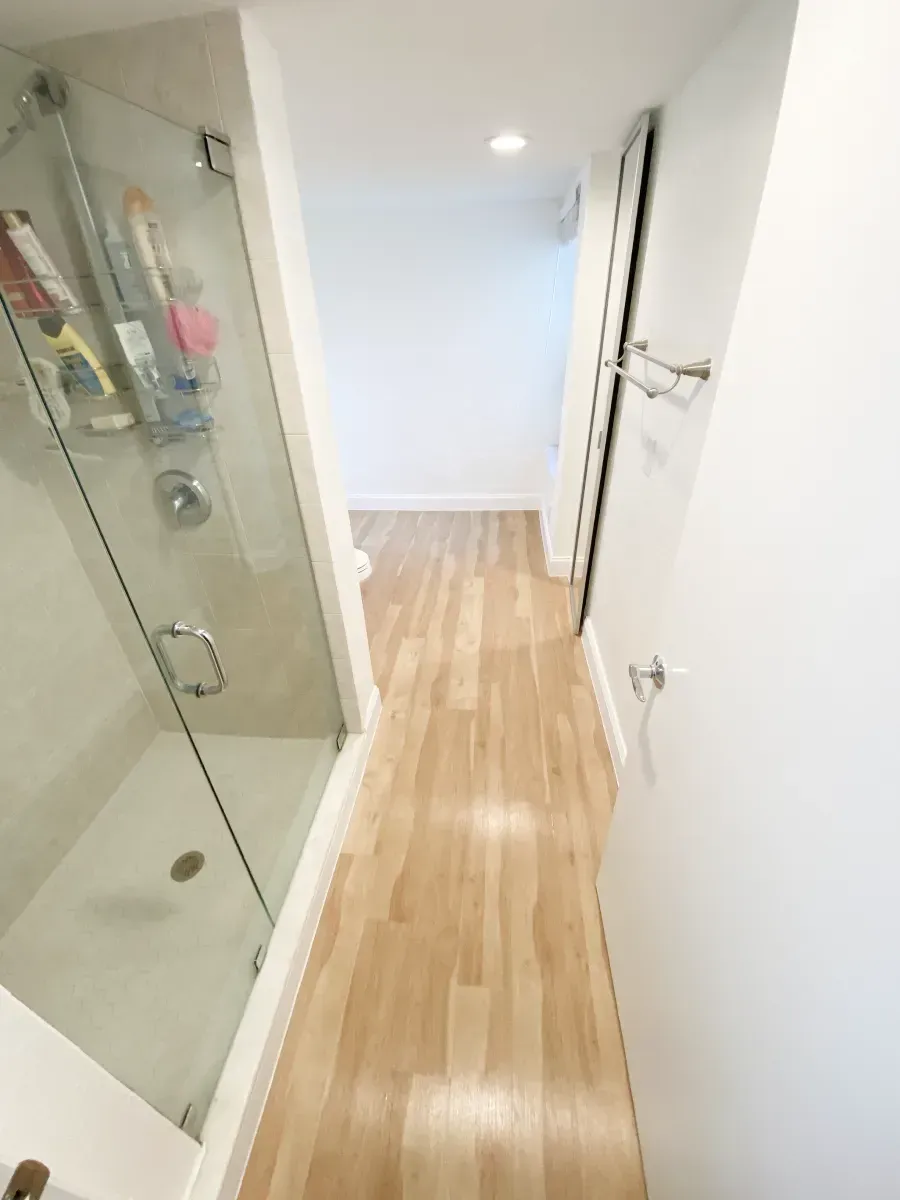 A brightly lit hallway with wood flooring, featuring a glass-enclosed shower on the left and white walls throughout.