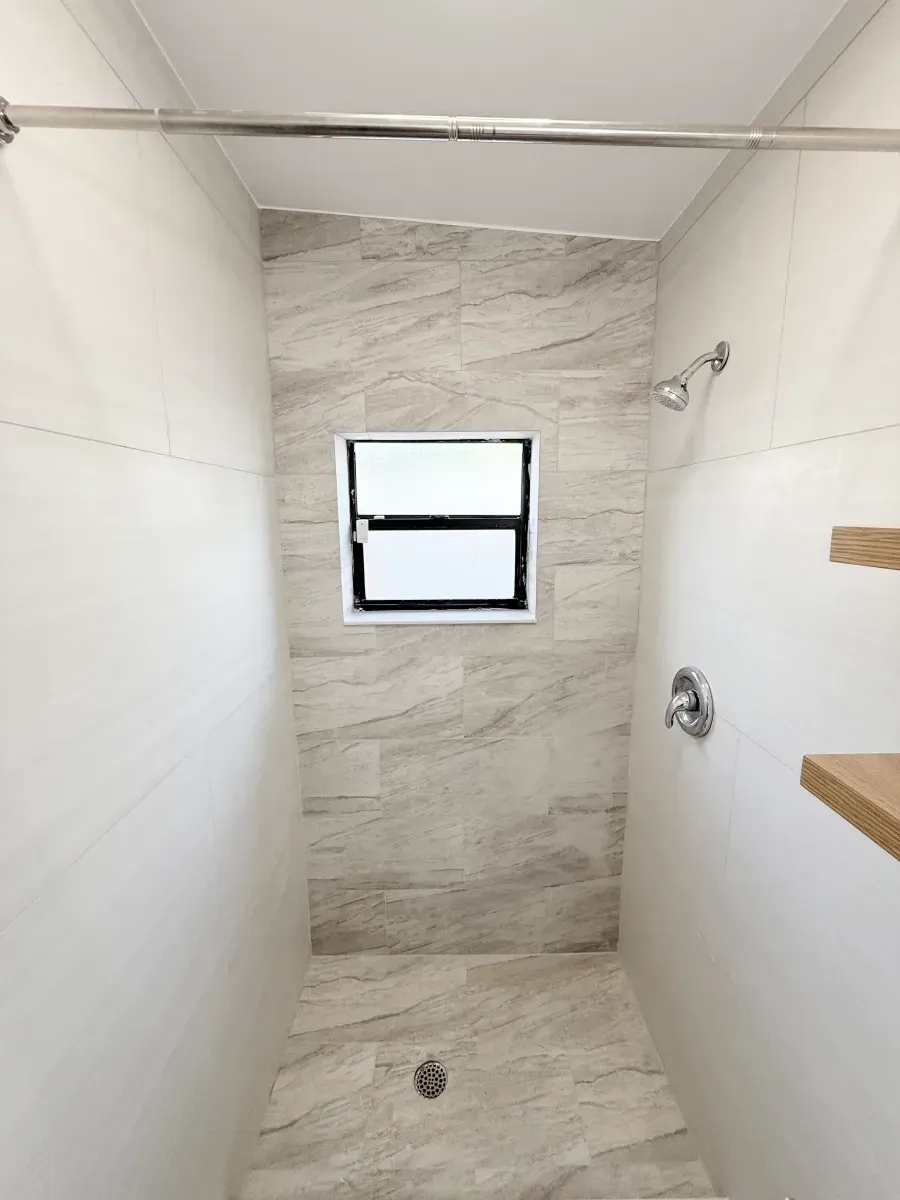 A shower stall with beige, wood-look tiled walls and floor, a small center window, and a silver shower head and faucet.