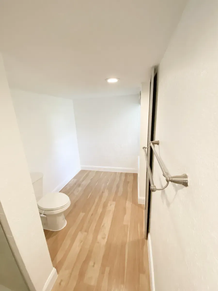 A view of a minimalist bathroom with a white toilet, light wood flooring, white walls, and a silver towel bar.