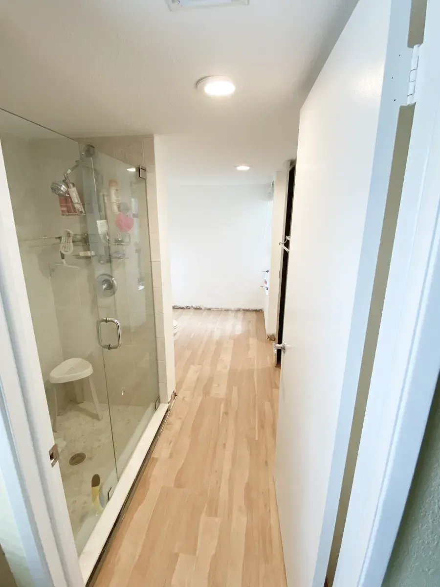 A narrow hallway leading to a bright room, featuring a glass-enclosed shower stall on the left and light wood flooring.