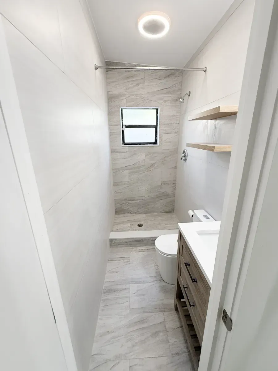 A narrow, modern bathroom with light-toned marble-look tile, a corner shower, a vanity, and two wooden wall shelves.