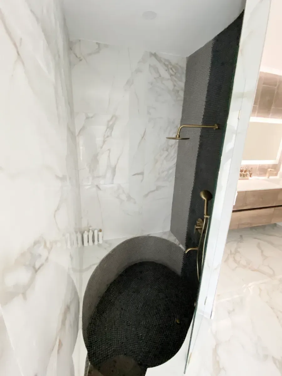 A modern shower with white marble walls, a dark stone floor, and gold fixtures, set within a bright bathroom.