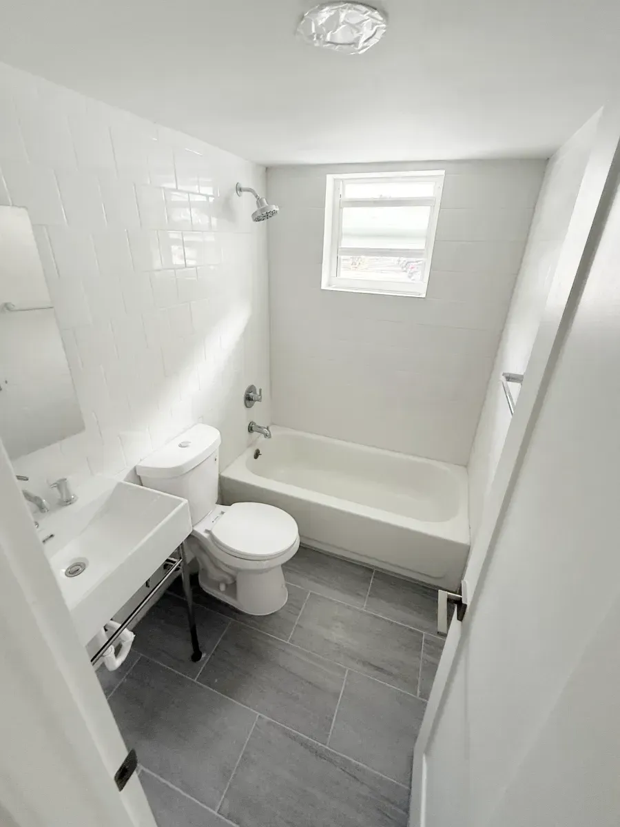 A small, bright bathroom with white walls, a pedestal sink, a white toilet, a bathtub with a showerhead, and gray tile floors.