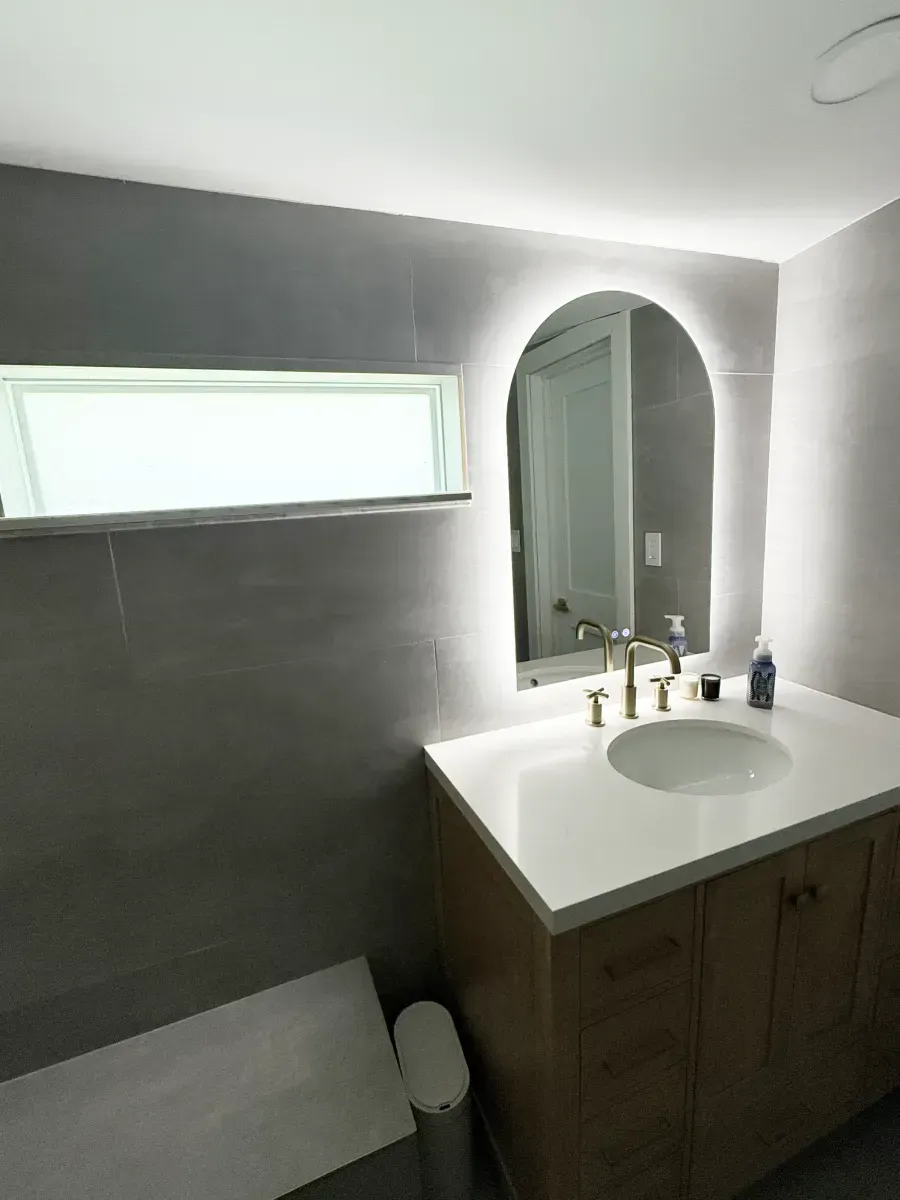 A modern bathroom with a wooden vanity, a white countertop, an arched LED-lit mirror, and grey tiled walls with a window.