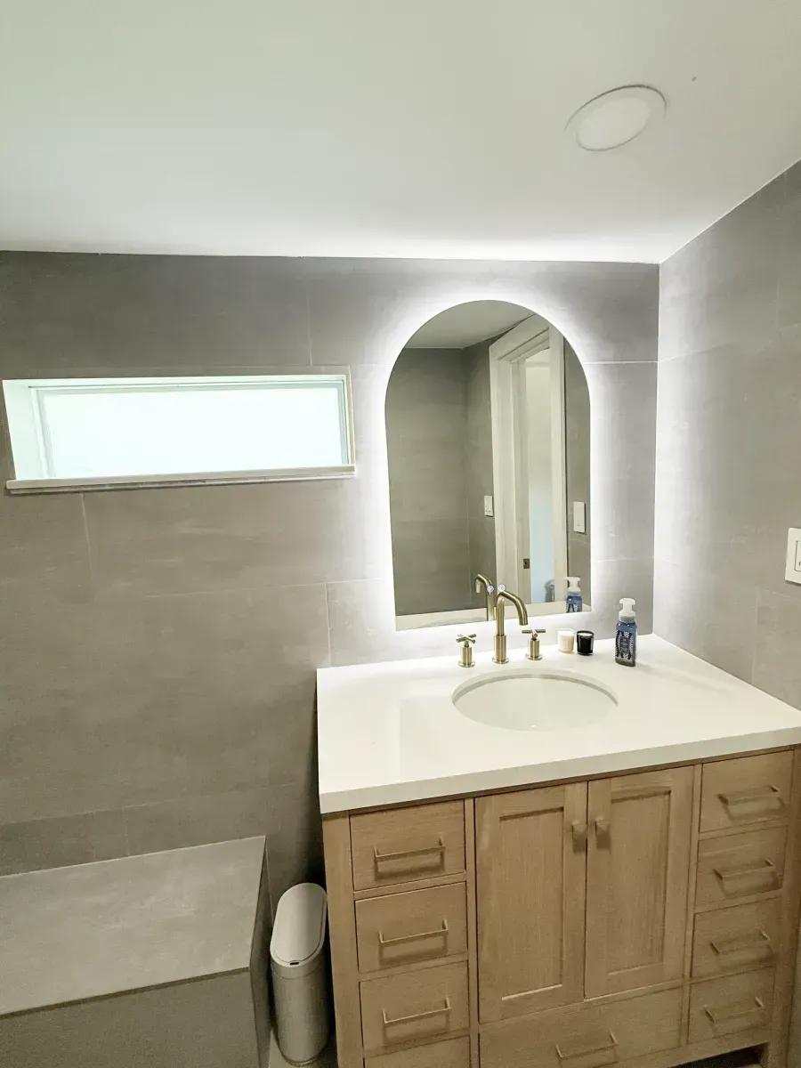 A bathroom vanity with a light wood cabinet, white countertop, sink, and an arched, LED-backlit mirror against gray tiles.