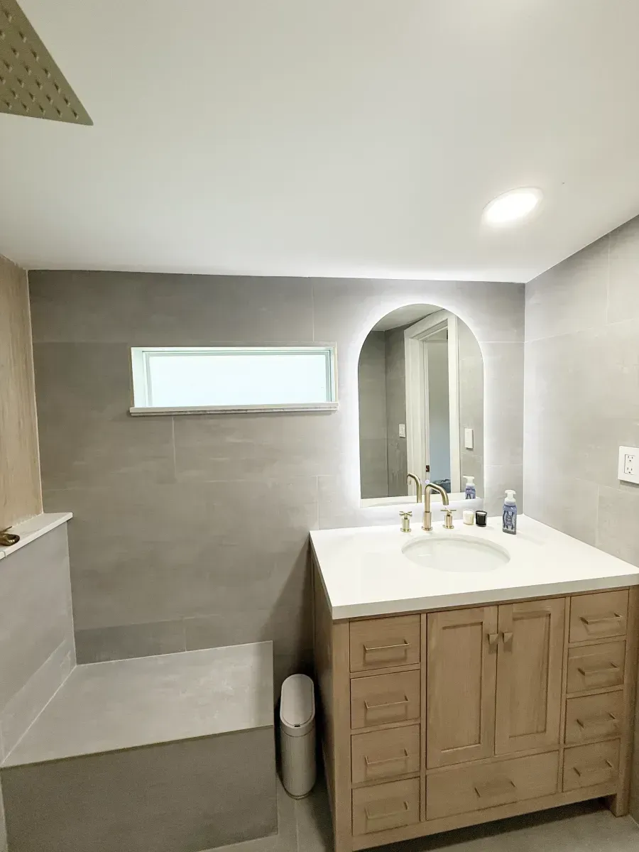 A bathroom with a gray-tiled shower bench, a wooden vanity with a white countertop, and an arched, backlit mirror.
