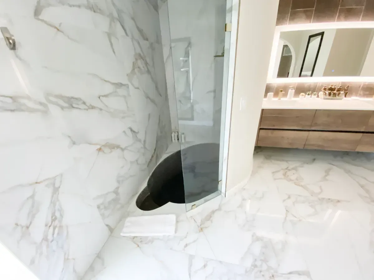 A bathroom with marble tile floors and walls, featuring a glass-enclosed shower and a vanity with an illuminated mirror.