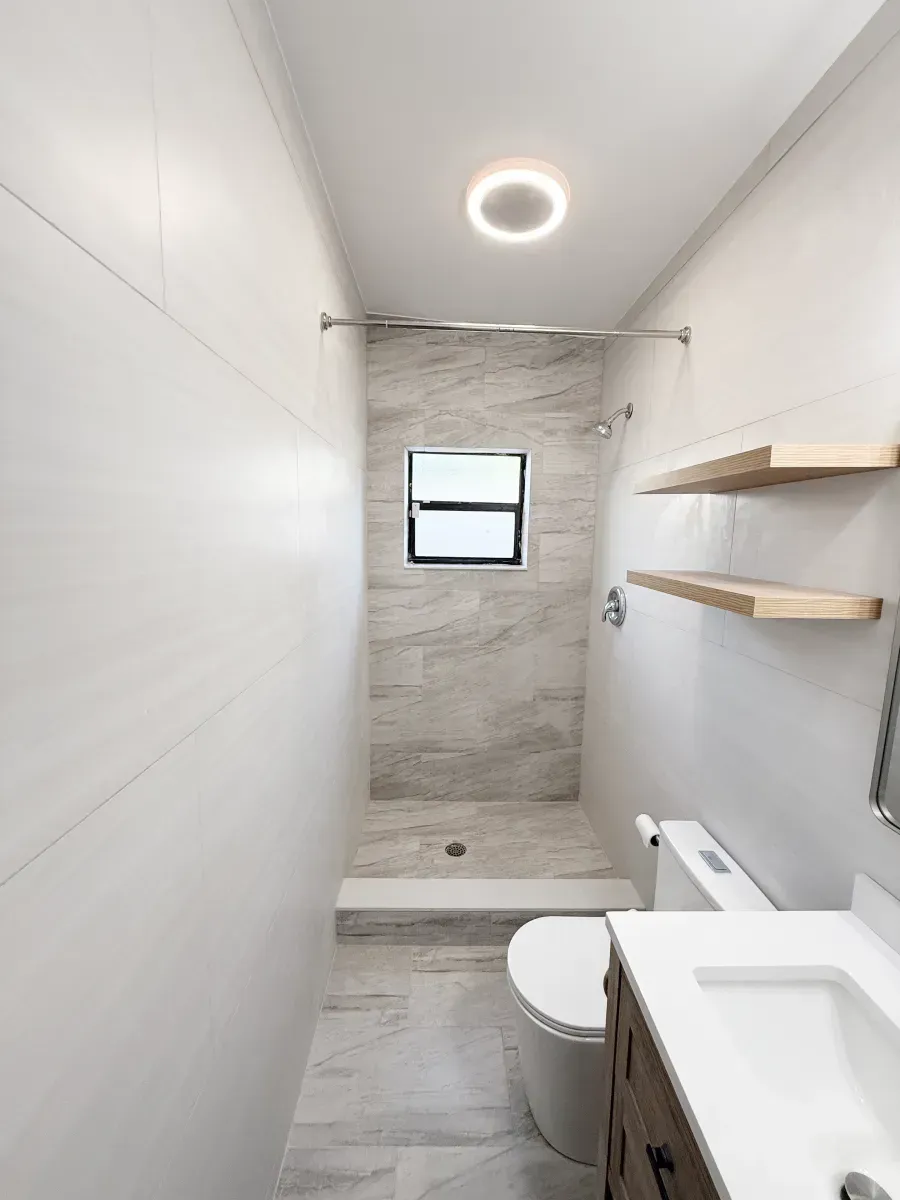 Modern bathroom with white walls, a wood vanity, floating shelves, and a walk-in shower with gray-toned tile flooring.