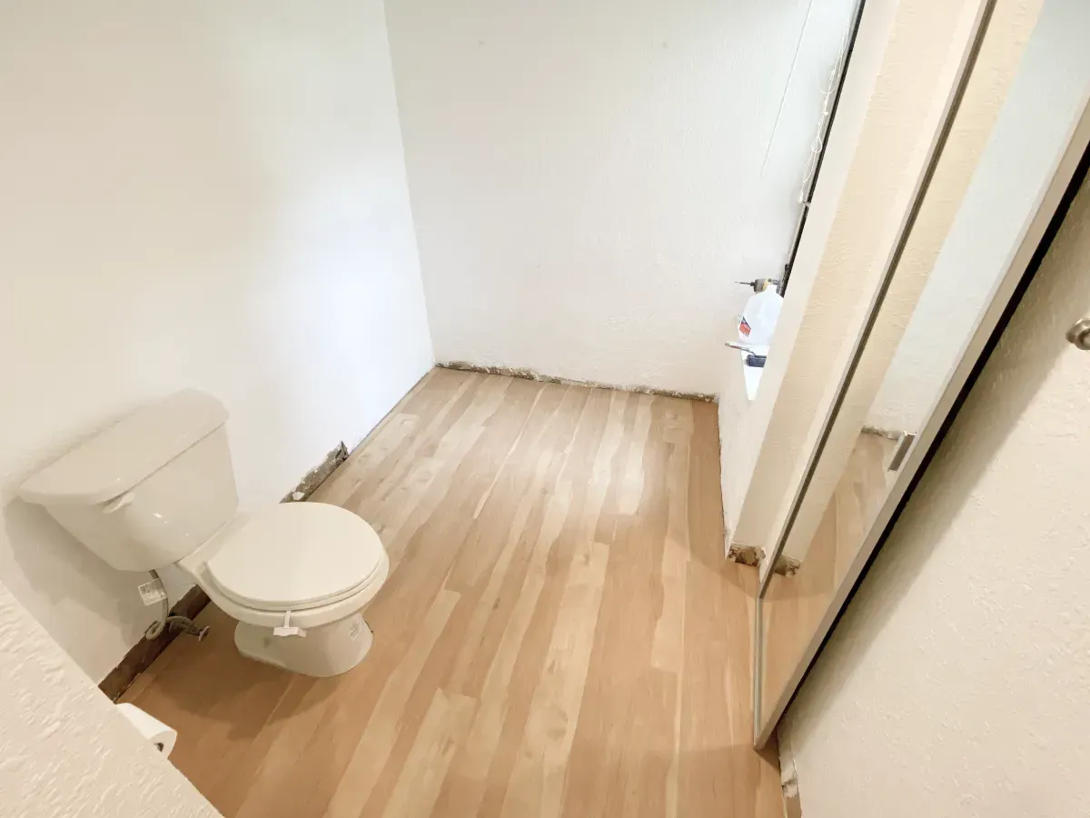 A white toilet sits in a small room with light-colored wood-look flooring and white walls, viewed from a high angle.
