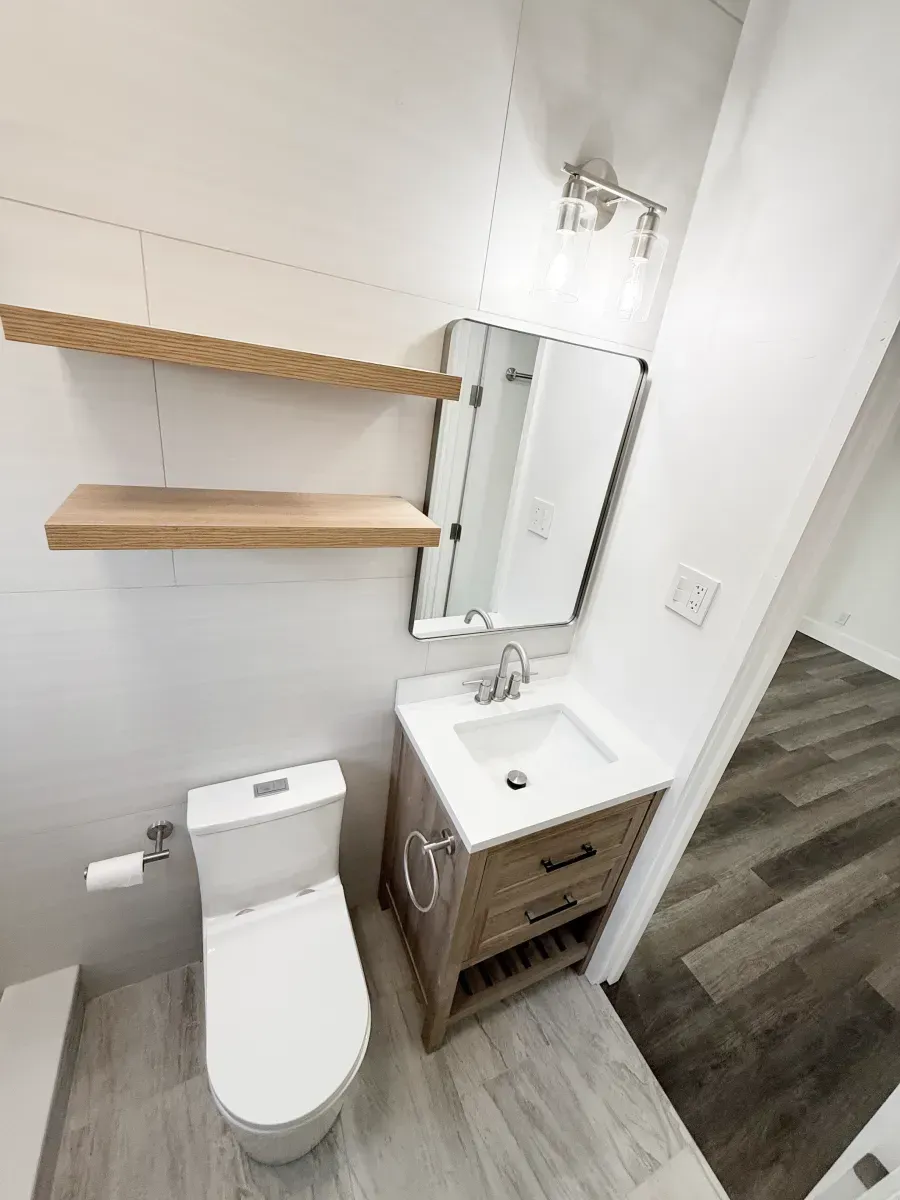 A modern bathroom with a white toilet, a wooden vanity with a white sink, and two wooden floating shelves on a white wall.