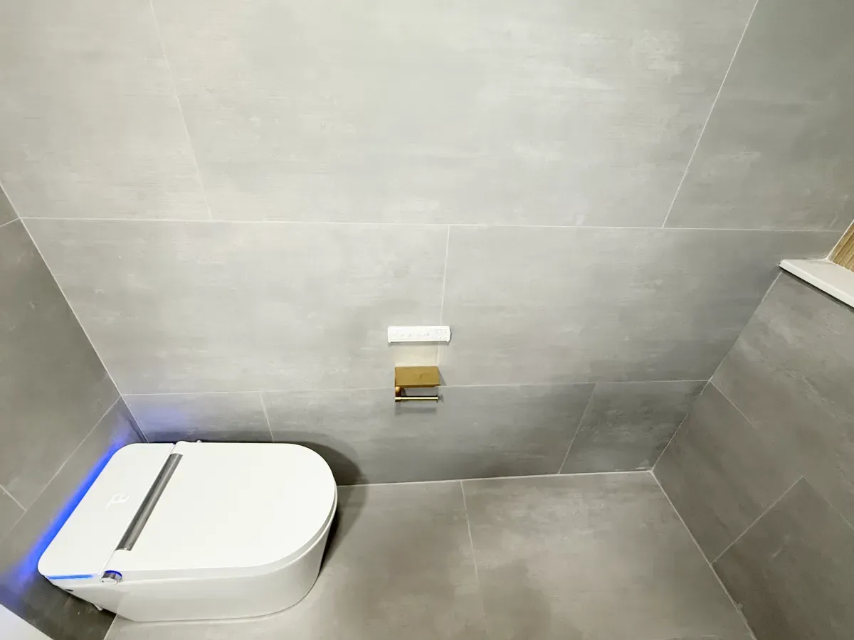 A white smart toilet with a glowing blue light sits in a modern bathroom with grey tiled walls and a gold toilet paper holder.