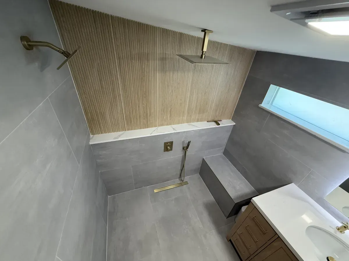 A modern bathroom shower area featuring gray tiled walls, a wood-slat accent wall, a rain showerhead, and a bench.
