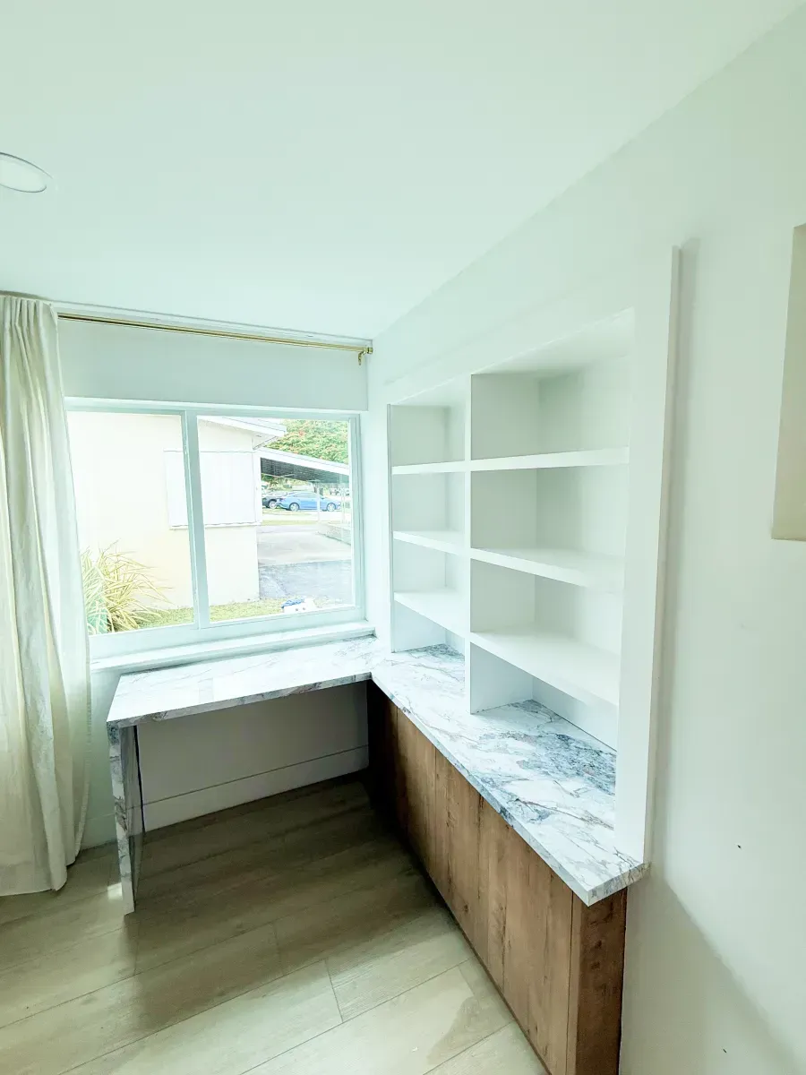 A white built-in desk and shelving unit with wooden base cabinets and a marble-patterned countertop by a bright window.