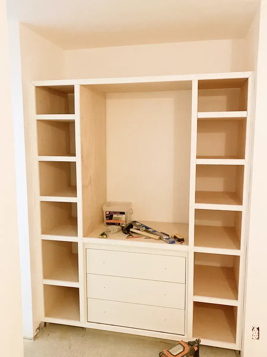 A custom white built-in shelving unit with drawers and open storage compartments, currently under construction in a room.