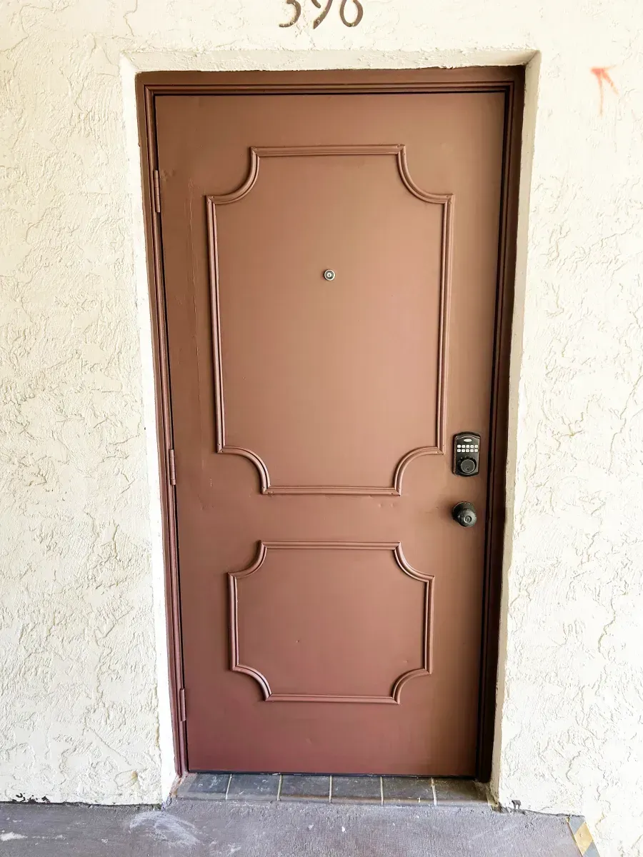 A brown front door numbered 398 with two rectangular molded panels, a door viewer, and a digital lock, set in a beige wall.