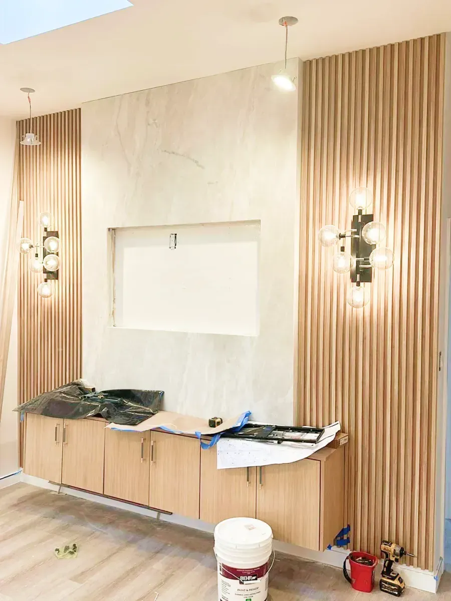 A modern interior wall with wood-slat panels, marble-style center, wall sconces, and floating cabinetry under construction.