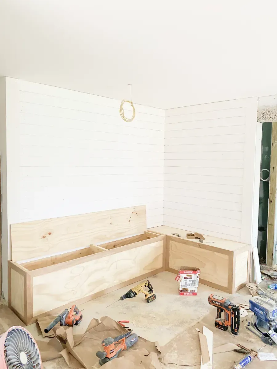 An L-shaped wooden bench seat under construction in a room with white shiplap walls and various tools on the floor.