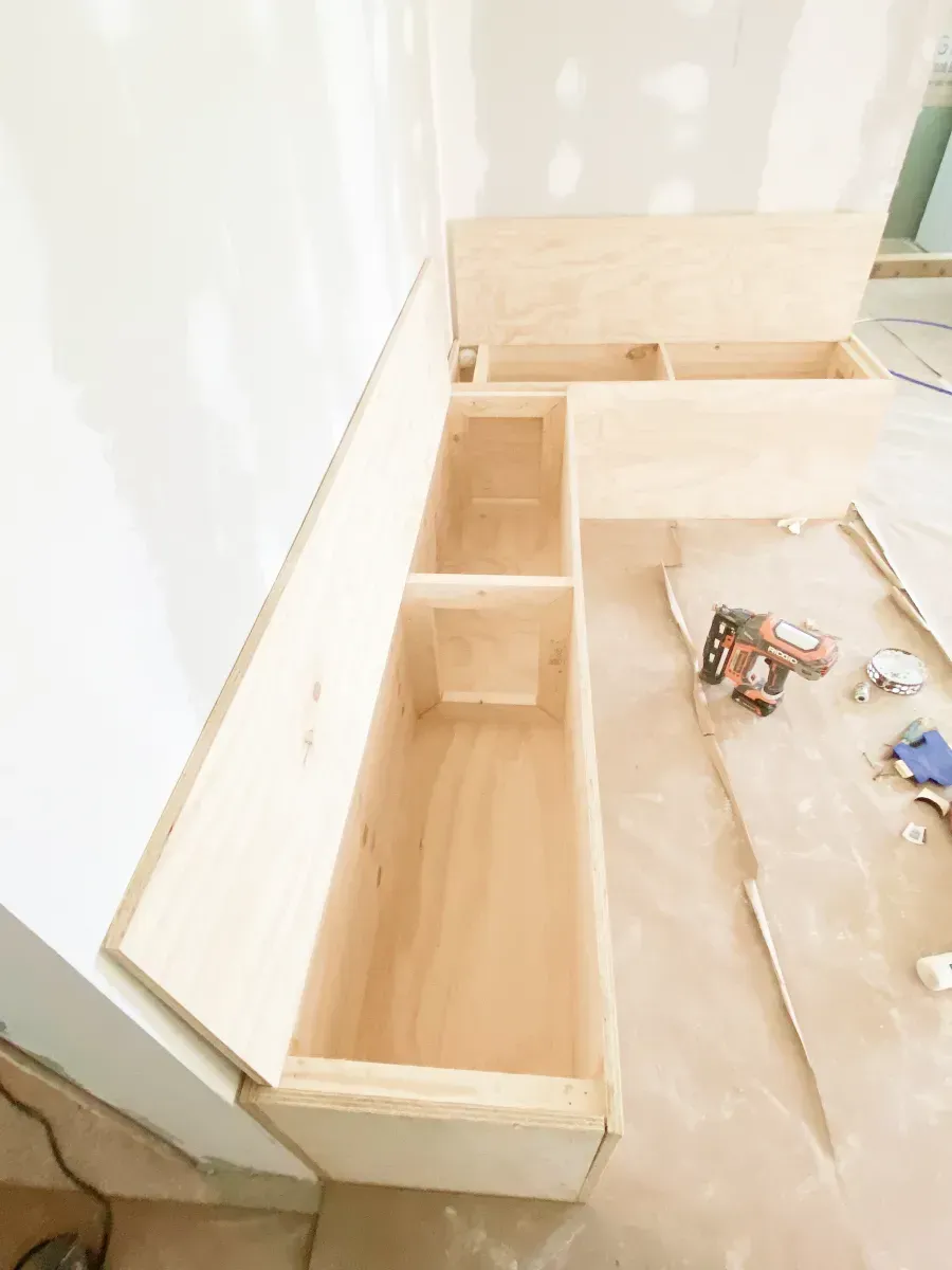 L-shaped wooden built-in bench under construction in a room with unfinished drywall and a nail gun on the floor.