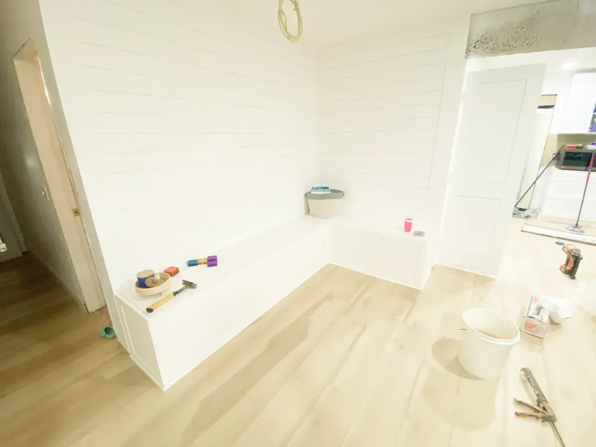 An L-shaped white built-in bench in a bright, unfinished room with light wood-look flooring.