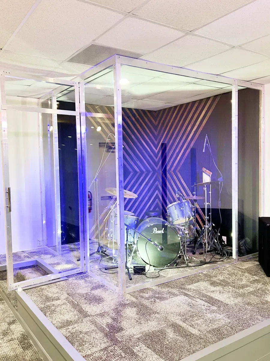 A Pearl drum kit inside a glass sound isolation booth on a carpeted platform in a room with patterned walls and blue light.