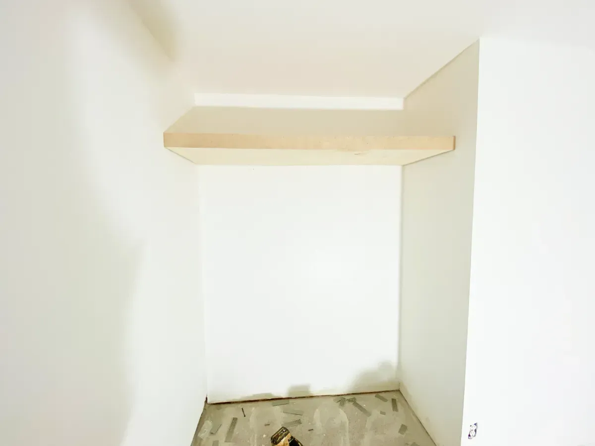 An unfinished white closet space featuring a single, light-colored wooden floating shelf installed near the ceiling.