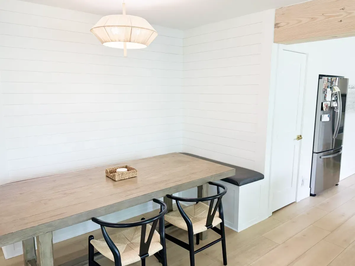 Dining area with a wooden table, two black chairs, a white bench, and a modern pendant light against a white brick wall.