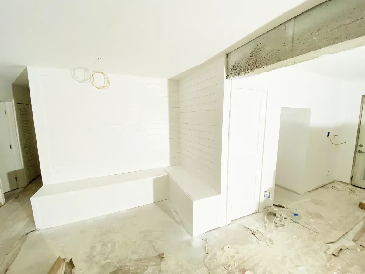 An interior view of a room under construction with white walls, a built-in bench, and exposed ceiling beams.
