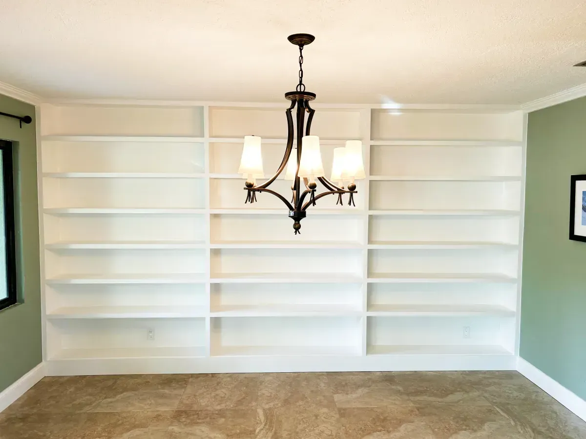 A wall of built-in white shelves with a hanging chandelier in front, flanked by sage green walls and tiled flooring.