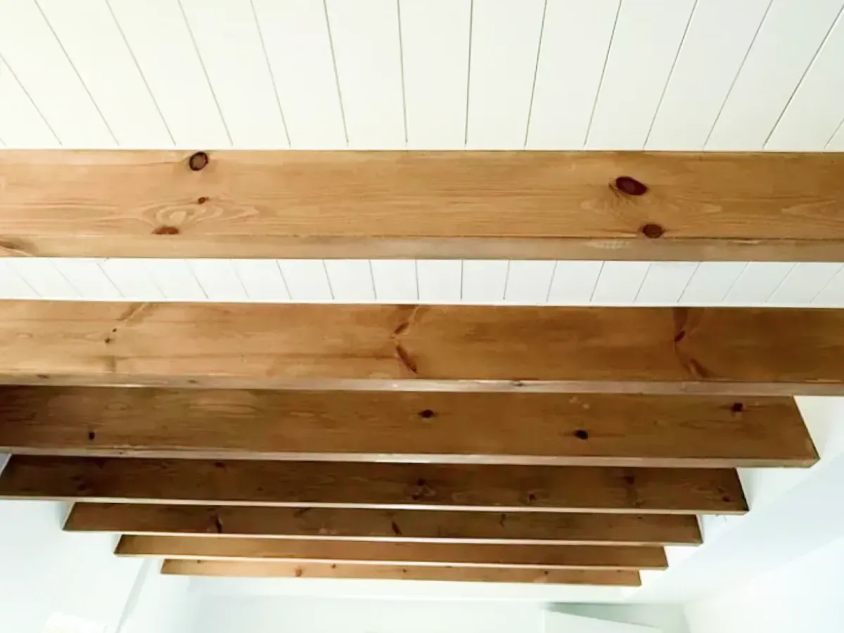 A low-angle view of a white paneled ceiling featuring several parallel, rustic wooden beams running across it.