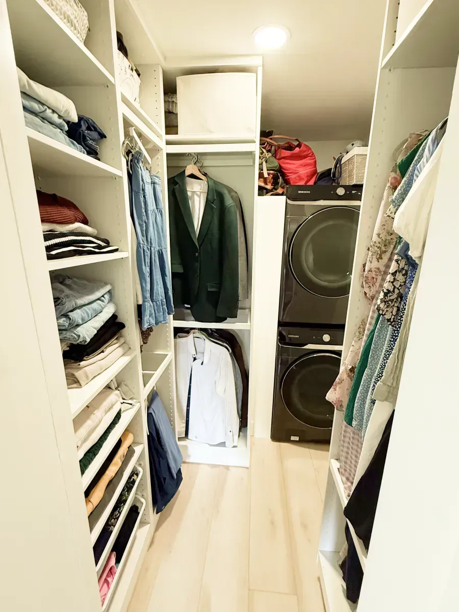 A walk-in closet with shelves of folded clothes, hanging garments, and a stacked washer and dryer at the far end.
