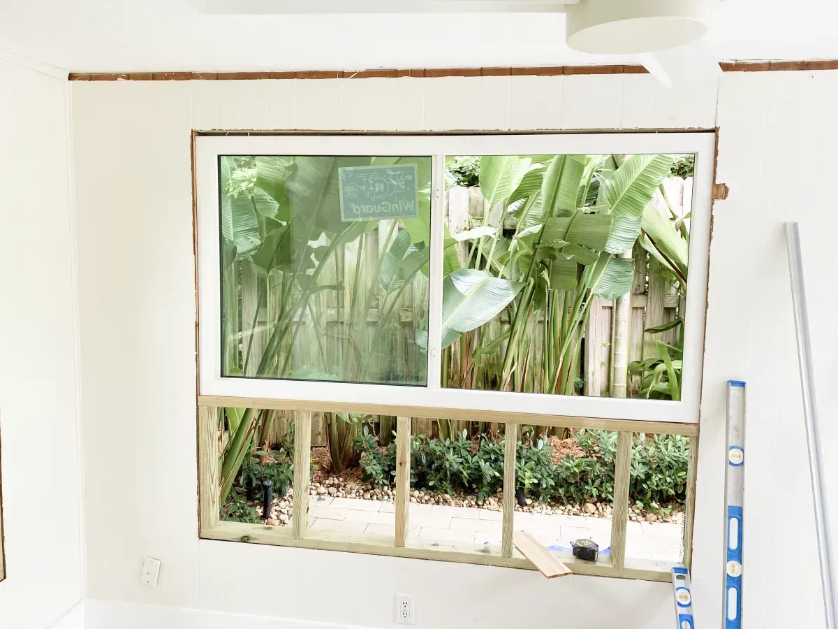 A window installed in a framed wall section during home renovation, showing green plants outside.