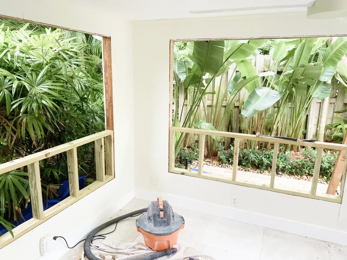 Two large window openings in an interior wall reveal lush green foliage outside, with framing installed below the openings.
