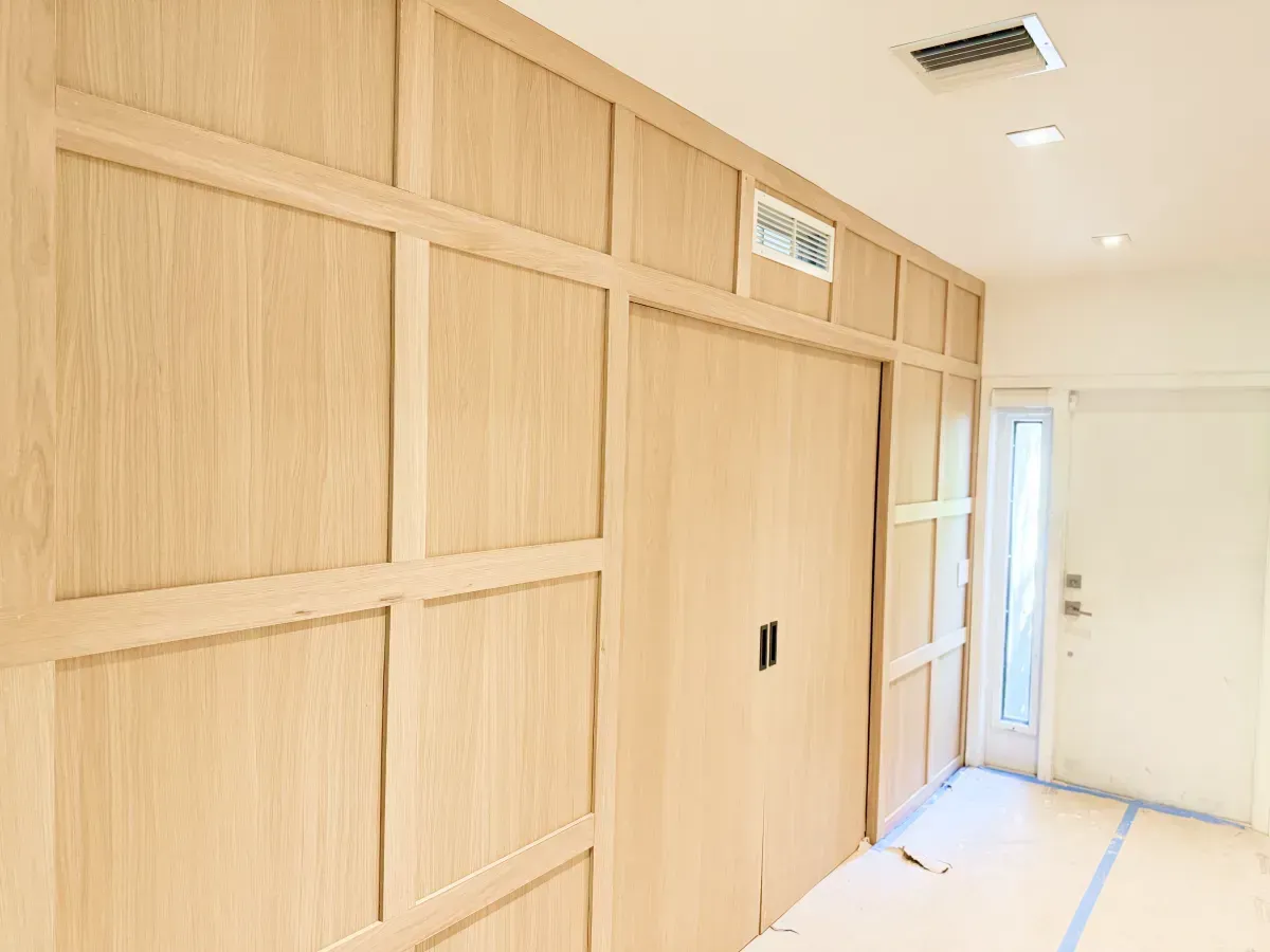 A light-wood paneled wall with a central sliding door, located in an interior hallway with white flooring.