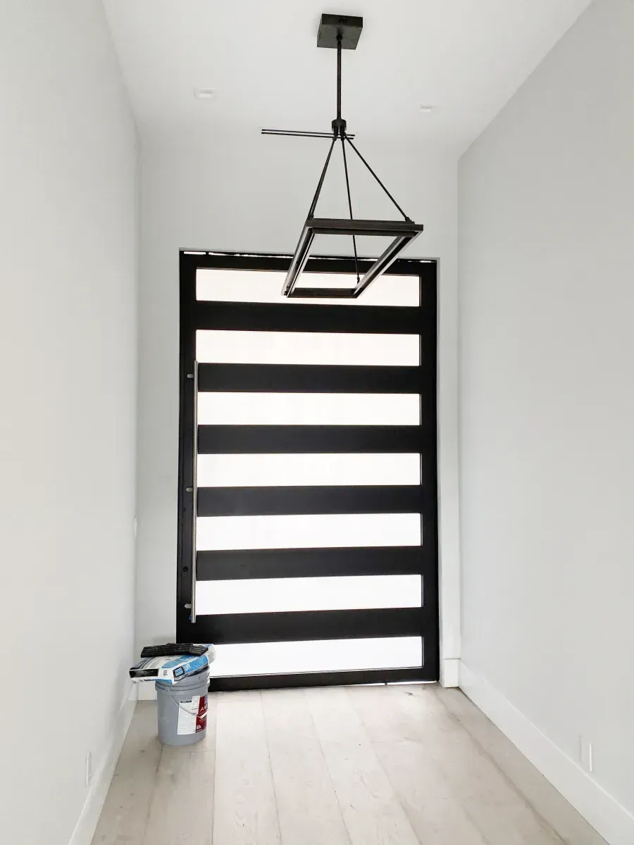 A narrow hallway with white walls and light wood floors, featuring a modern black front door with glass horizontal panels.