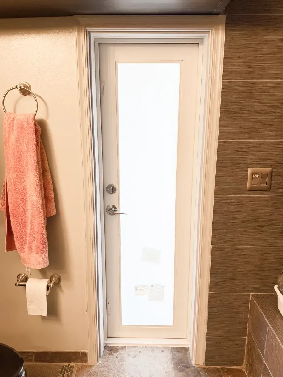 A white door with a frosted glass panel centered in a room with light and dark brown tiled walls and a pink towel.