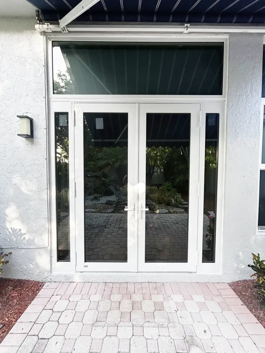 A set of white-framed double glass doors with side lights and a transom window, set in a light grey stucco building.