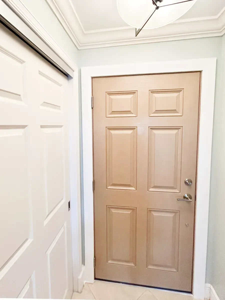A six-panel tan door set in a white frame next to a white closet door in a hallway with a light fixture above.