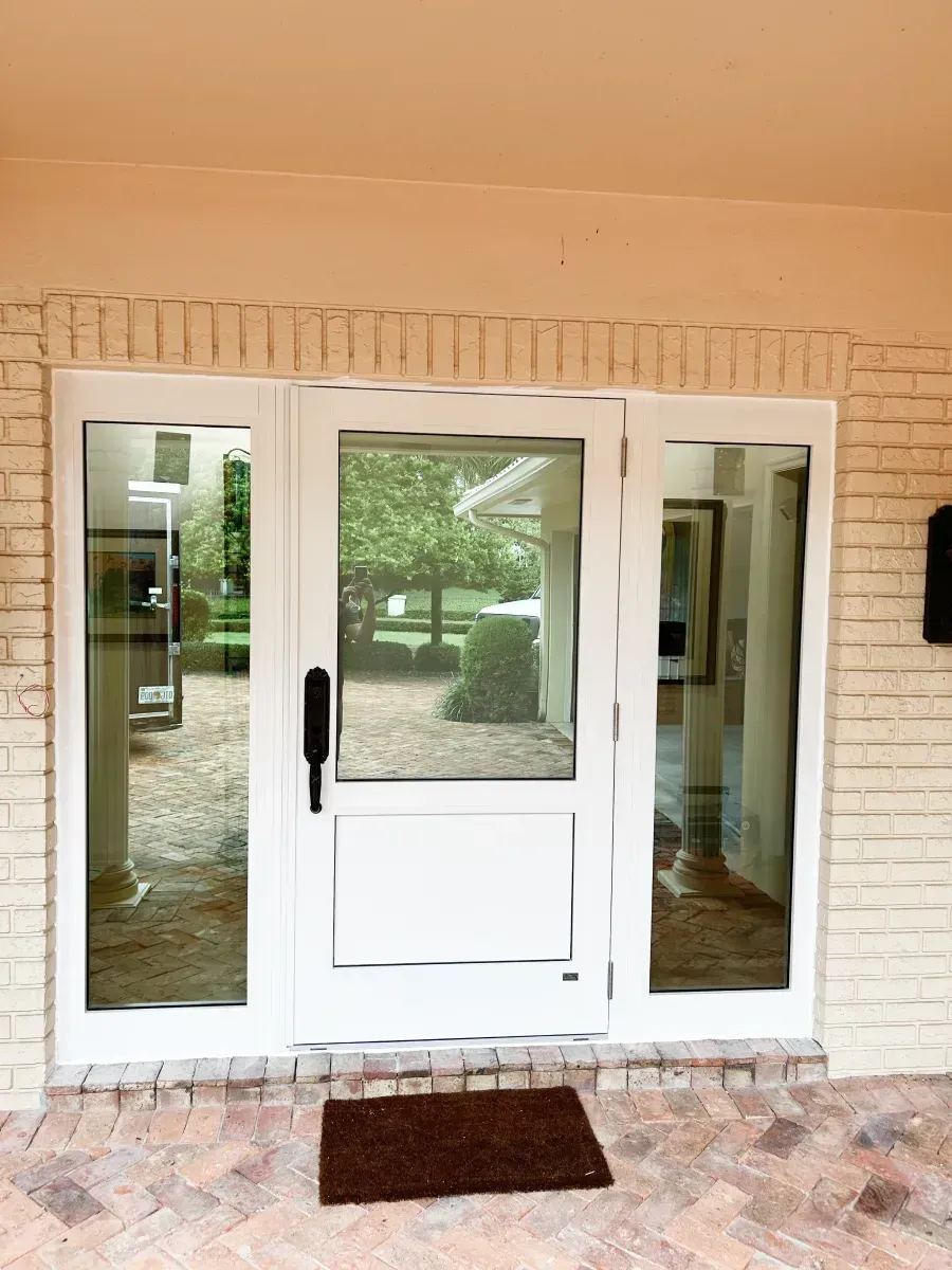 A white front door with glass side panels set into a beige brick wall, featuring a dark handle and a brown door mat.