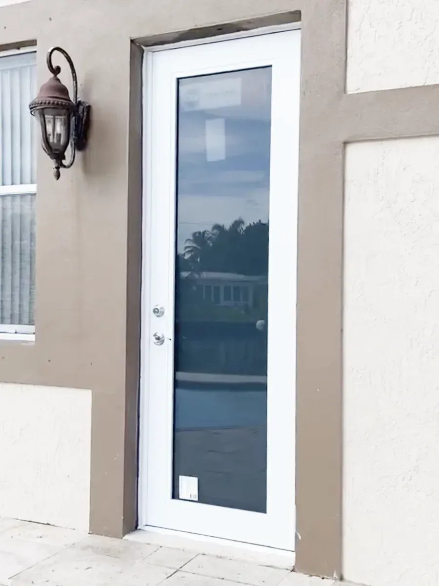 A white door with a long glass panel set into a tan, stucco-style exterior wall next to a wall-mounted lantern light.
