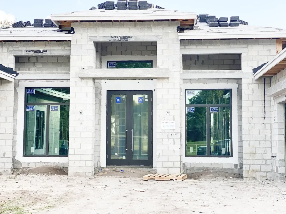 Front view of a house under construction with concrete block walls, installed dark-framed windows, and double doors.